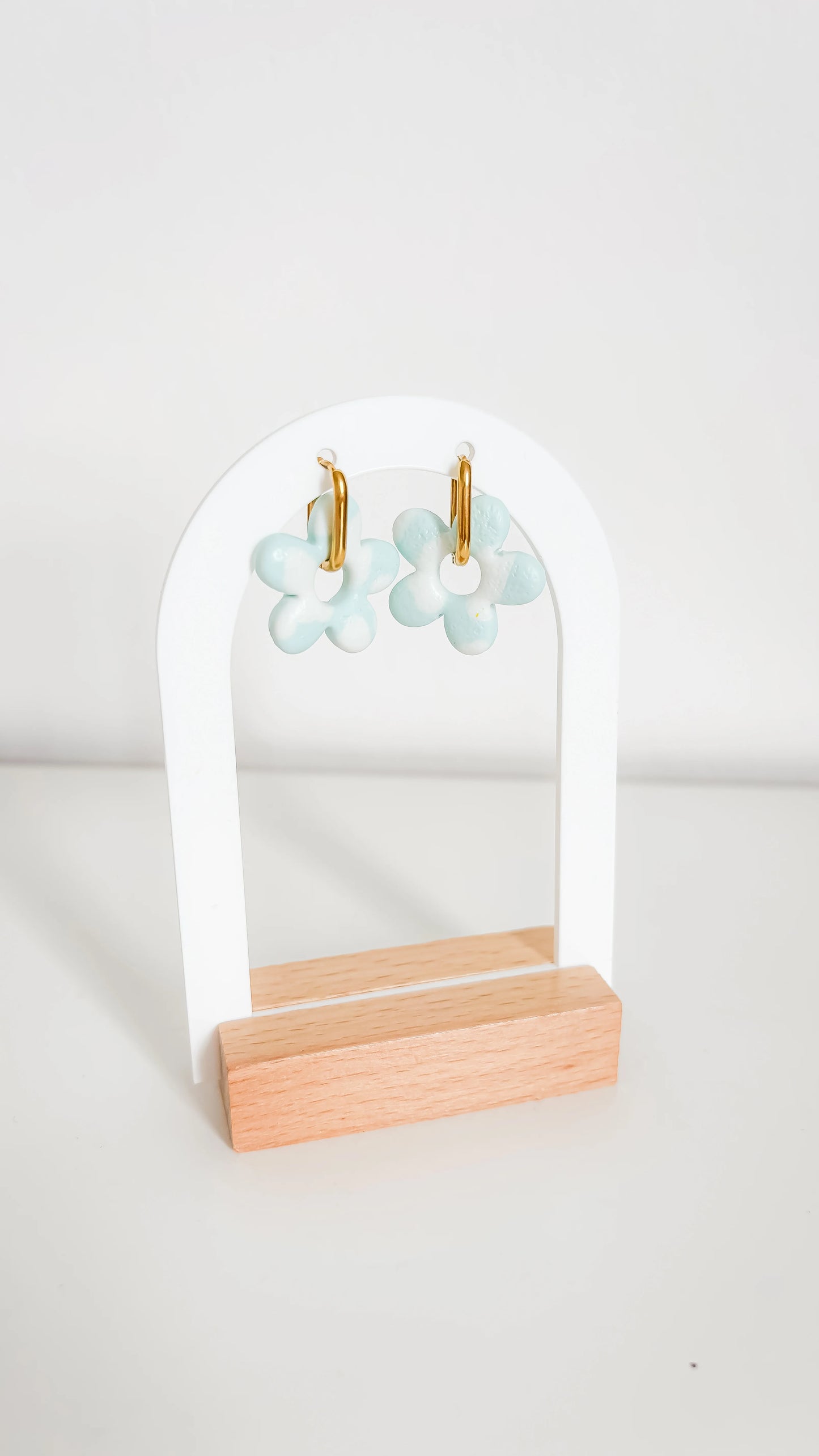 Boucles d’oreilles fleuries bleu clair sur support doré, suspendues à un présentoir moderne en bois clair.