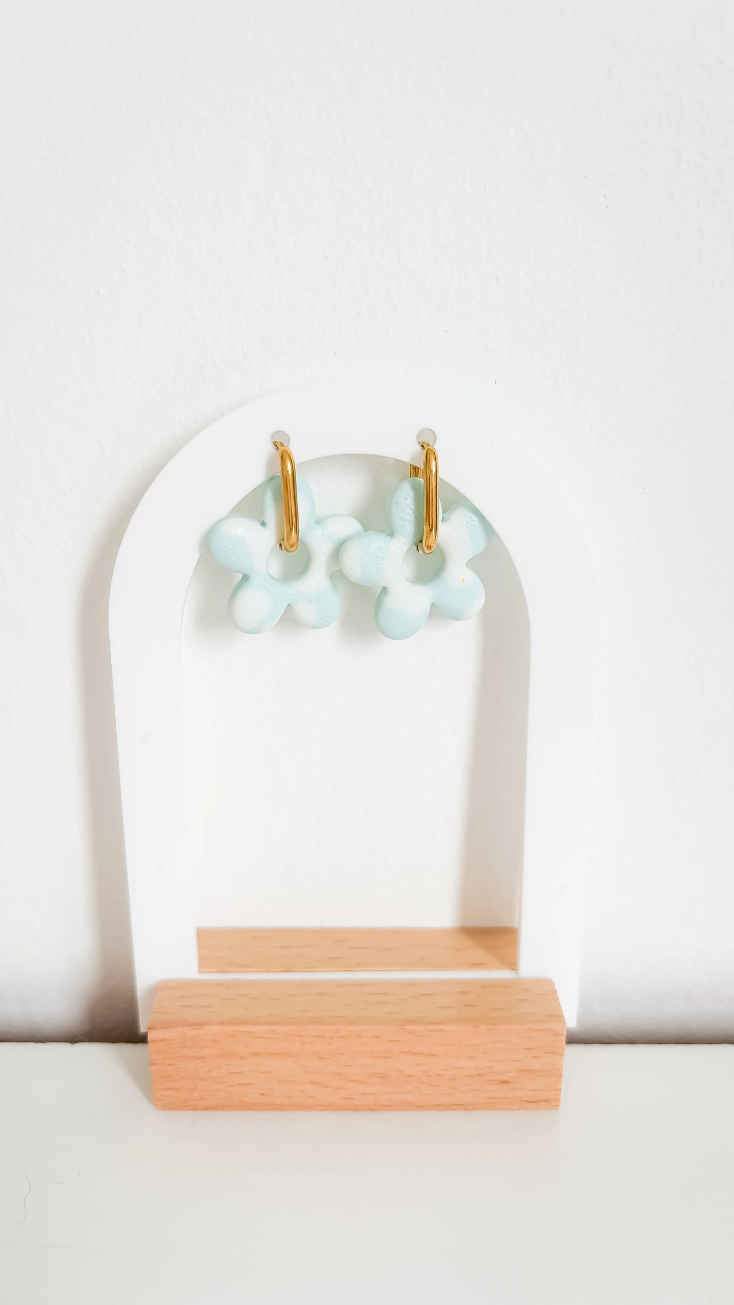 Créoles dorées avec fleurs en pâte polymère bleu ciel et blanc, suspendues sur un présentoir minimaliste.