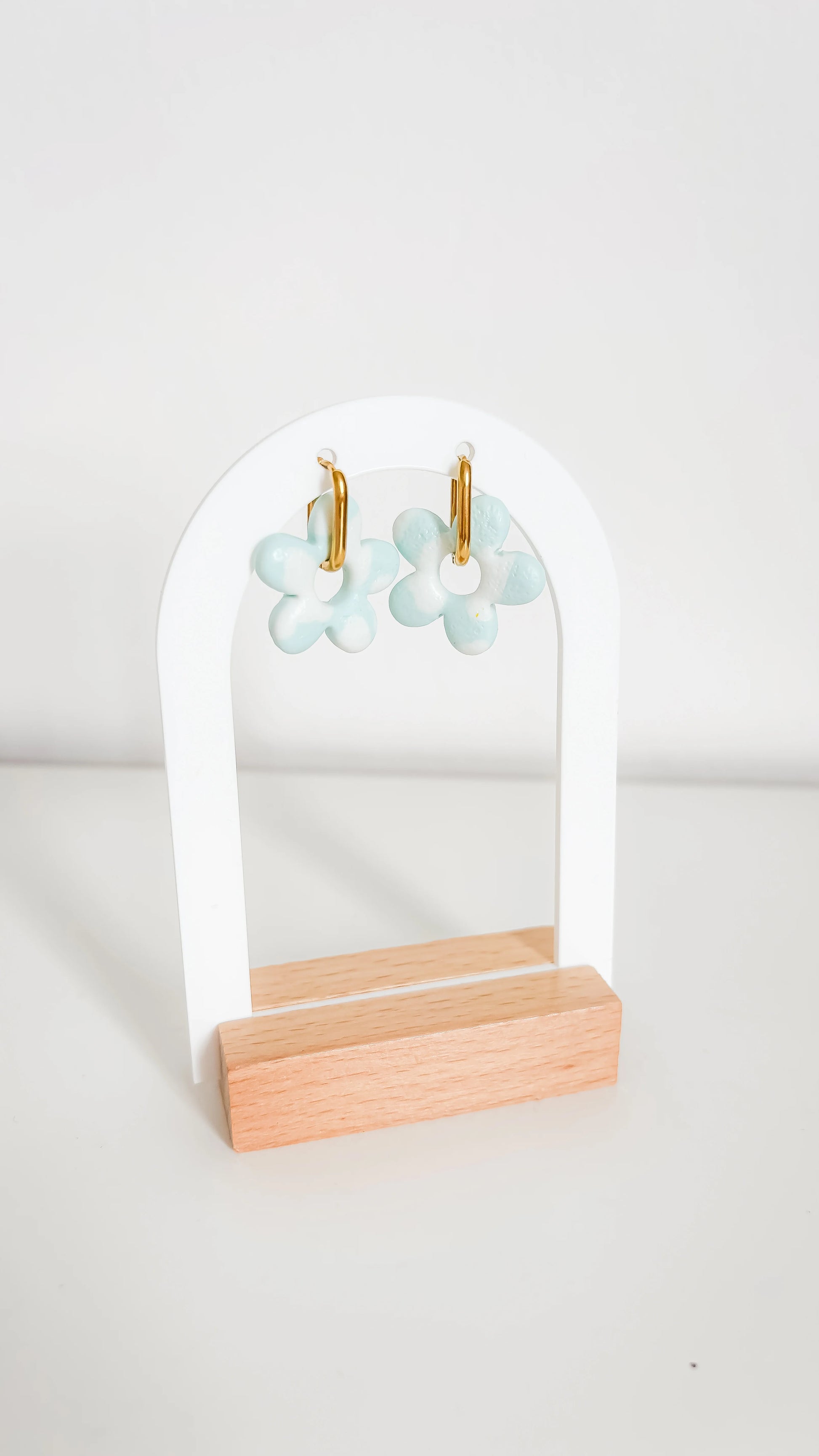 Boucles d’oreilles fleuries bleu clair sur support doré, suspendues à un présentoir moderne en bois clair.
