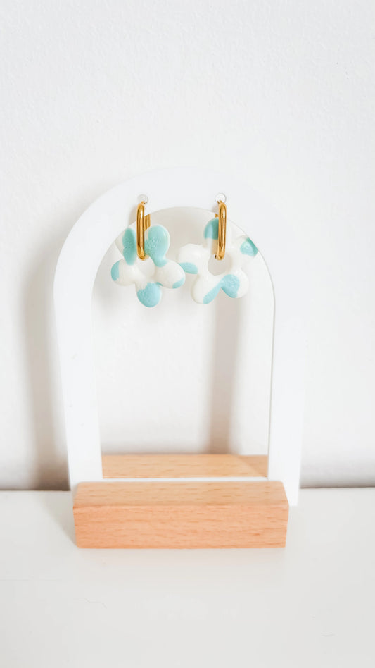 Boucles d’oreilles en forme de fleurs bleu pastel et doré, montées sur un support blanc et bois pour mise en valeur.